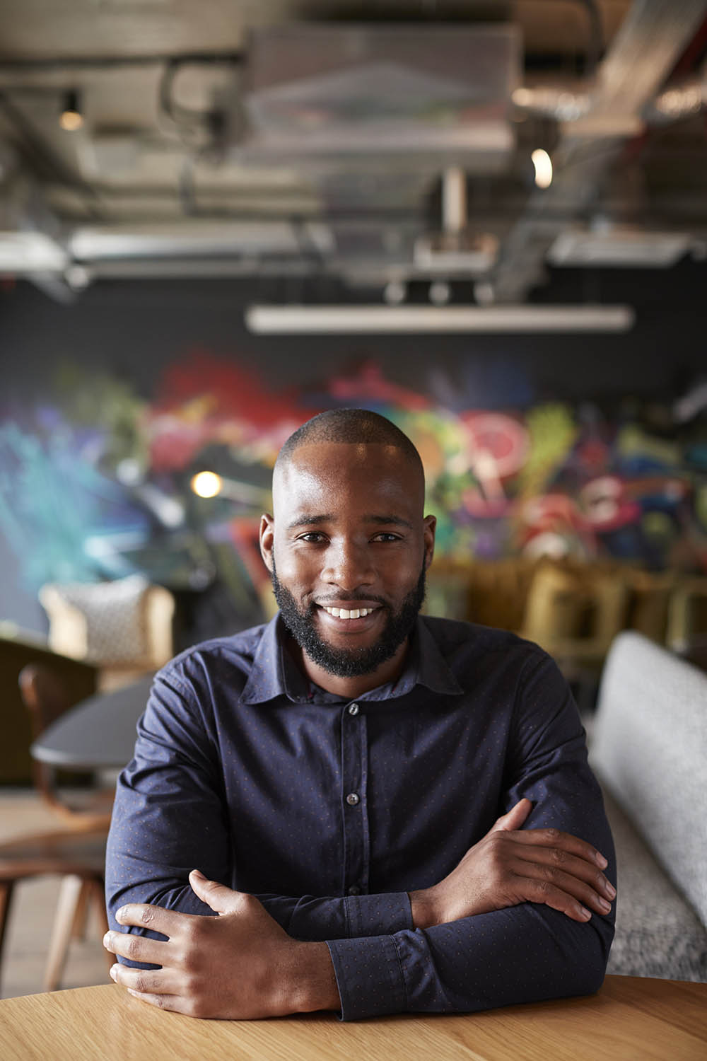 Mid adult black male creative sitting at a table with arms crossed smiling to camera, close up Venture Group